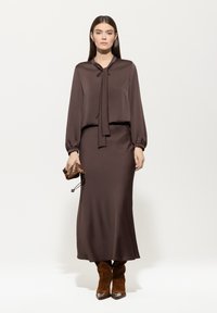 Braune Satin-Bluse mit Krawattenkragen und elastischen Manschetten, kombiniert mit einem passenden braunen, bodenlangen Rock; weiche Textur. Braune Wildleder-Stiefel bis zum Knie.