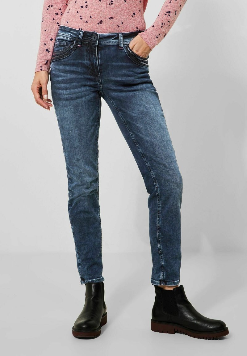 Jeans de mezclilla azul oscuro con corte recto, que presentan un diseño clásico de cinco bolsillos y un ligero desgaste. Combinados con botas negras de tobillo.