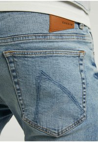 Jasnoniebieskie dżinsy z denimu z charakterystycznym wzorem na tylnej kieszeni, jasnobrązową skórzaną naszywką z napisem "CHASIN'" oraz pomarańczowymi przeszyciami.