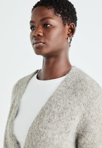 Cardigan deschis în față, din lână texturată gri deschis, asortat cu un tricou alb cu guler rotund. Detalii subtile și croială relaxată.