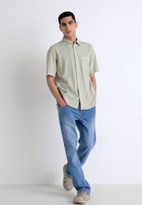 Chemise boutons court à manches courtes de couleur vert clair avec une poche sur la poitrine gauche, associée à un jean bleu clair et des baskets beiges, sur un fond neutre.