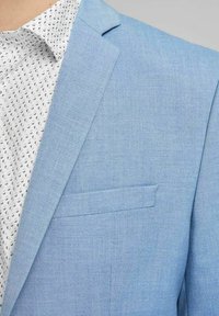 Ljusblå blazer i texturerat tyg, med en enda bröstficka och notchad krage, kombinerad med en vit skjorta med svart mönster.