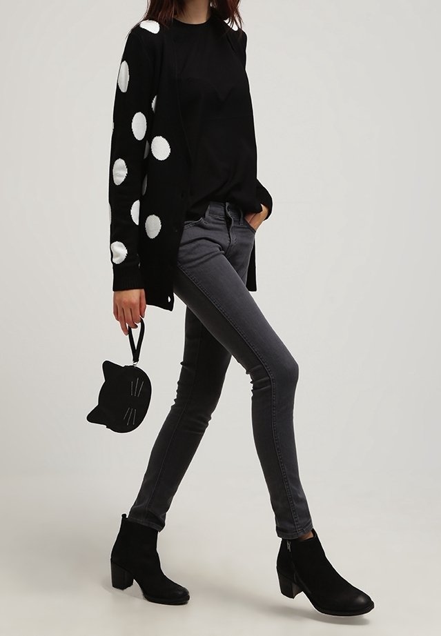 Femme portant un cardigan noir à pois, un haut noir, un jean skinny gris foncé, des bottines noires, tenant un petit sac noir en forme de chat.