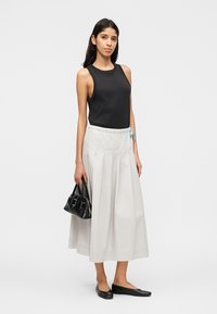 Γυναίκα με μαύρο τοπ χωρίς μανίκια, λευκή μίντι φούστα, μαύρες φλατ παπούτσια, κρατώντας μικρή μαύρη τσάντα, μπροστά από απλό φόντο.