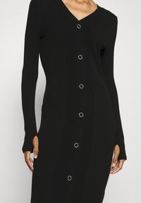 Robe en maille côtelée noire à manches longues, col en V, et fermetures à boutons sur le devant ornés de garnitures circulaires argentées ; fentes latérales près des poignets.