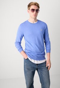 Helles blaues Strickpullover mit langen Ärmeln und rundem Halsausschnitt, versehen mit einem kleinen gelben Logo. Kombiniert mit blauen Jeans und Sonnenbrille.