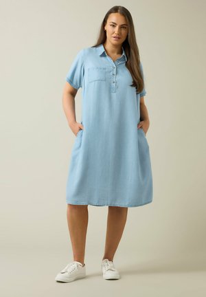 Femme debout les mains dans les poches, portant une robe boutonnée bleu clair longueur genoux et des baskets blanches, sur un fond beige.
