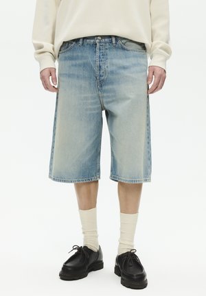 Osoba ubrana w jasno-niebieskie dżinsowe szorty do kolan, kremową koszulę z długim rękawem, białe skarpetki i czarne skórzane buty sznurowane.