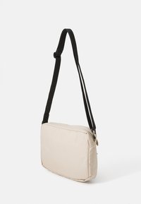 Beige crossbody taske med et blødt stof, rektangulær form og justerbar sort rem; har en lynlåslukning og ingen udvendige lommer.