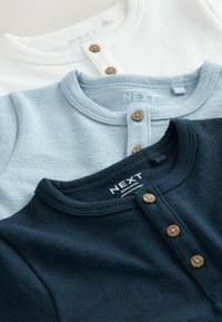 Három texturált henley póló egymásra rakva: egy fehér, egy világoskék és egy tengerészkék, mindegyik kerek nyakkivágással és fa gombokkal elöl.