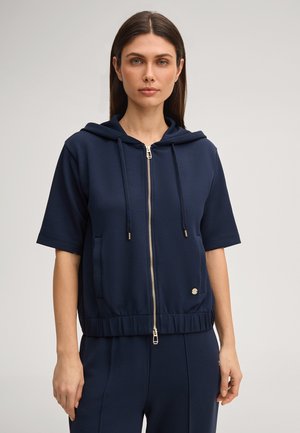 Donna che indossa una giacca blu navy con cappuccio, maniche corte e zip, con coulisse, abbinata a pantaloni blu navy, in piedi contro uno sfondo semplice.