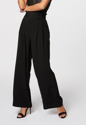 Kobieta w czarnych spodniach z wysokim stanem i szerokimi nogawkami, z guzikami w pasie oraz czarnych sandałach na wysokim obcasie, z założonymi skrzyżowanymi ramionami.