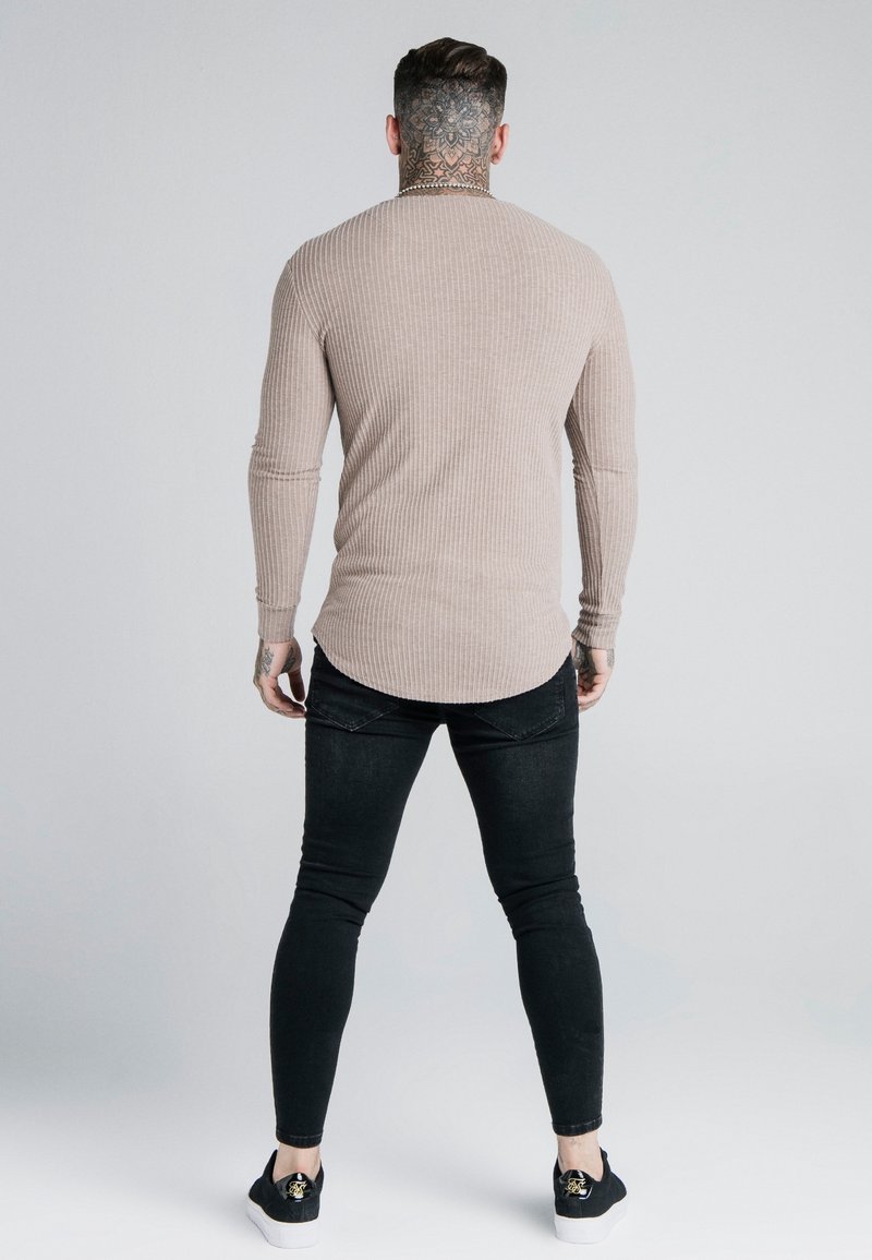 Muž s tetováním na krku, oblečený v béžovém žebrovaném tričku s dlouhým rukávem, černých slim kalhotách a černých teniskách, stojí zády k fotoaparátu.