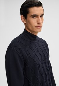Navyblauwe gebreide trui met een hoge ribbelkraag en een textuurpatroon met diamantvormen en verhoogde accenten door het gehele kledingstuk.