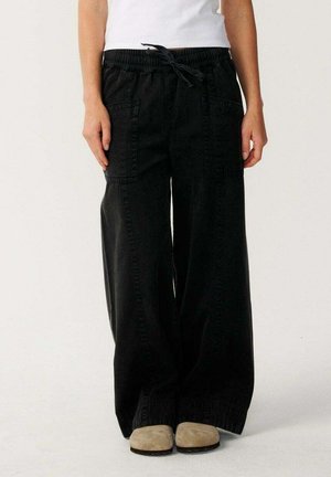 Personne portant un pantalon noir à jambes larges avec poches plaquées devant, un haut blanc et des chaussures beiges, debout devant un fond clair.