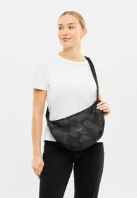 Geantă neagră pentru umăr, realizată dintr-un material texturat cu un model subtil. Prezintă o formă curbată și o curea ajustabilă. Logo-ul este expus vizibil.