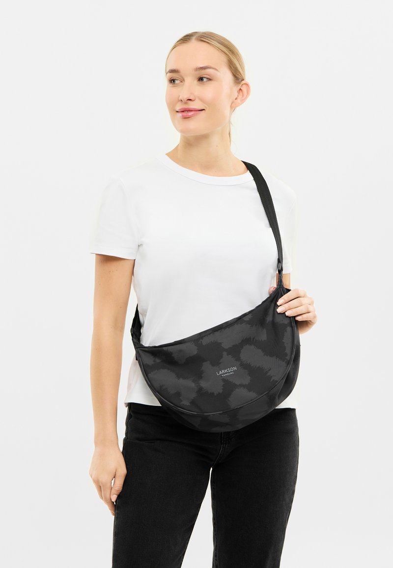 Geantă neagră pentru umăr, realizată dintr-un material texturat cu un model subtil. Prezintă o formă curbată și o curea ajustabilă. Logo-ul este expus vizibil.