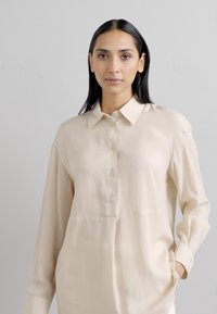 Camicia lunga beige chiaro con colletto. Presenta una chiusura con bottoni sul davanti e tasche laterali. Texture liscia con vestibilità comoda.