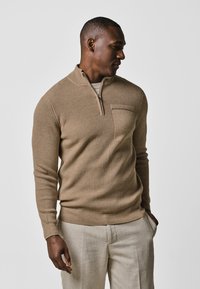 Beige gestrickter Pullover mit Rippstruktur, einem Reißverschlusskragen und einer Brusttasche. Getragen mit hellen Hosen. Enganliegend und strukturiert.