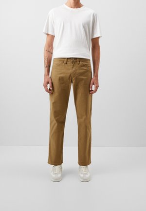 Homme debout portant un t-shirt blanc à manches courtes, pantalon marron clair droit, baskets blanches, sur un fond uni gris clair.