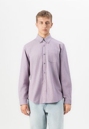 Young man wearing a long-sleeve lavender button-up shirt and light blue jeans standing against a white background.