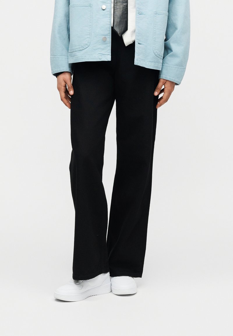 Persoon die zwarte broek met wijde pijpen, witte sneakers, lichtblauwe jas, wit shirt en zwarte stropdas draagt tegen een effen achtergrond.