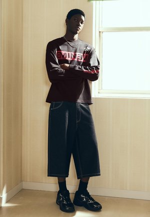 Mladý muž so založenými rukami, oblečený v čiernom tričku Eminem s dlhým rukávom, široké rifľové šortky, čierne tenisky, stojaci pri okne v interiéri.