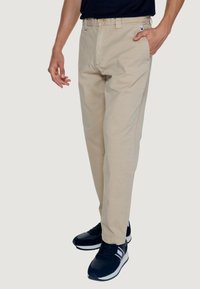 Pantaloni chino beige con taglio a gamba dritta, chiusura con bottoni e tasche laterali, abbinati a sneaker blu navy con dettagli bianchi e rossi.