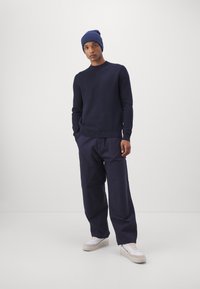 Marineblå sweater og beanie, løstsiddende mørke bukser og hvide sneakers. Glat tekstur og enkelt, funktionelt design.