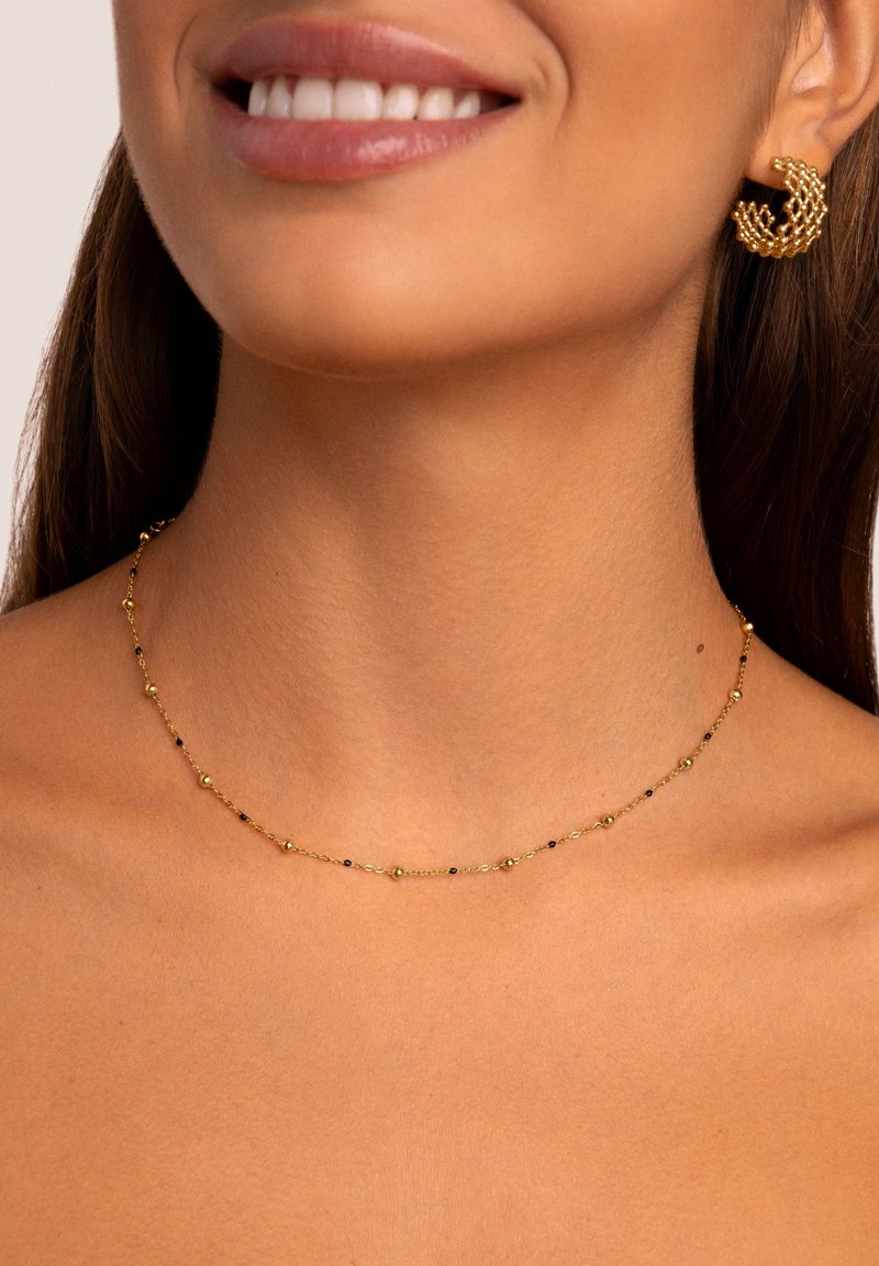 Collana d'oro con piccole perle rotonde e accenti neri, indossata con un orecchino ad anello in oro texturizzato, su uno sfondo neutro.