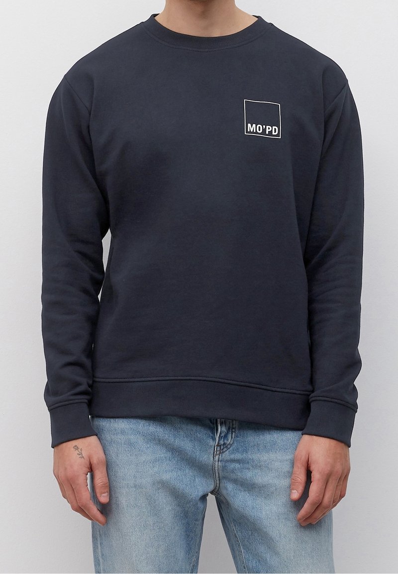 Homme portant un sweat-shirt bleu marine avec un carré contour blanc et le texte "MO'PD" sur la poitrine gauche, assorti à un jean bleu clair.