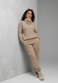 Beige knitted sweater with a cowl neck and chain detail, paired with beige cargo pants featuring multiple pockets and matching sneakers.