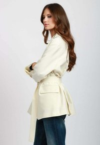 Giacca avorio con cintura dal design sartoriale, dotata di colletto strutturato, maniche lunghe e tasche laterali, abbinata a jeans blu.