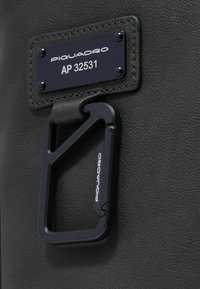 Produit en cuir noir avec une étiquette de marque en métal argenté portant l'inscription "PIQUADRO AP 32531" et un mousqueton noir attaché pour le détail de la quincaillerie.