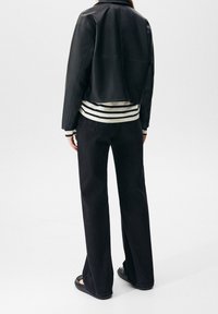 Personne debout, portant une veste noire courte, un jean large noir, une chemise à rayures à manches longues et des sandales noires, dos tourné à la caméra.