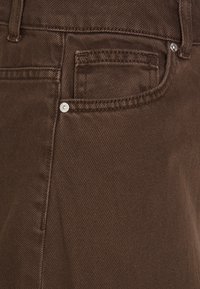 Pantalones de denim marrón con un acabado texturizado, un pequeño bolsillo frontal y acentos de hardware metálico plateado. Costuras visibles a lo largo de las costuras.