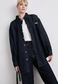 Mørk denim oversized skjorte med tradisjonell krage, hvite knappelukninger og en lomme på brystet. Komplementert med en enkel hvit topp.
