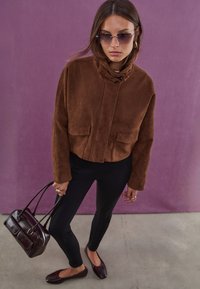 Giacca cropped in finta suede marrone con due tasche frontali, colletto rotondo e chiusura a morsetto. Pantaloni neri aderenti e borsa bordeaux.