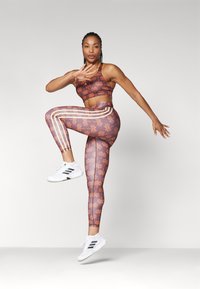 Sportig crop top och leggings-set i brunt med orange spindelmönster, accentuerat med vita ränder; parat med vita sportskor.