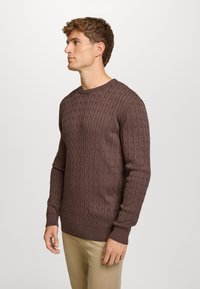 Brązowy sweter o splocie warkoczowym z okrągłym dekoltem, z prążkowanymi mankietami i dolnym obrzeżem, z fakturowanym wzorem, noszony na jasnych spodniach.