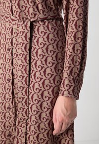 Abito a maniche lunghe in tessuto beige con un motivo a spirale color bordeaux, dotato di bottoni e una cintura coordinata in vita. Texture liscia.