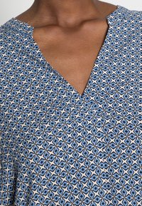 Nahaufnahme einer Person, die ein blaues und weißes geometrisches gemustertes Hemd mit V-Ausschnitt trägt.