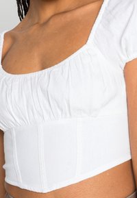 Vit avskuren topp med fyrkantig halsringning, korta puffärmar och samlad textur längs bodicen. Slitstark, mjuk tyg med synliga sömdetaljer.