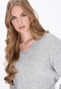 Hellgrauer, gestrickter Pullover mit V-Ausschnitt, der ein strukturiertes Muster aufweist. Weiches Material und lässige Passform mit dezentem Glanz.