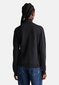 Dunkelgrauer Strickpullover mit hohem Kragen, weicher Textur und langen Ärmeln, kombiniert mit blauen Denim-Jeans, die ein grünes Etikett-Detail aufweisen.