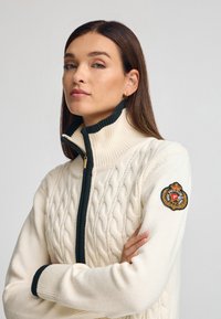 Kabelgebreide trui in crème met een hoge kraag, groene ritsdetails en een ronde patch met een wapenschild op de linkermouw.