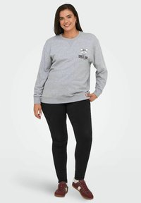 Felpa grigia con la scritta "SMILING" e grafica di un fiocco. Abbinata a pantaloni neri e sneakers rosse. Il modello si trova su uno sfondo neutro.