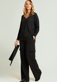 Czarny top z długim rękawem i dekoltem w serek o subtelnej fakturze, zestawiony z szerokimi, czarnymi spodniami typu cargo z bocznymi kieszeniami. Całość utrzymana w ciemnej kolorystyce.