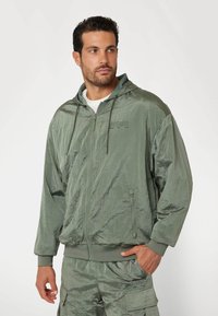 Vízálló zöld zipzáros kapucnis pulcsi texturált felülettel, húzózsinórral ellátott kapucnival és kenguru zsebekkel, összepárosítva megfelelő cargo sortokkal.