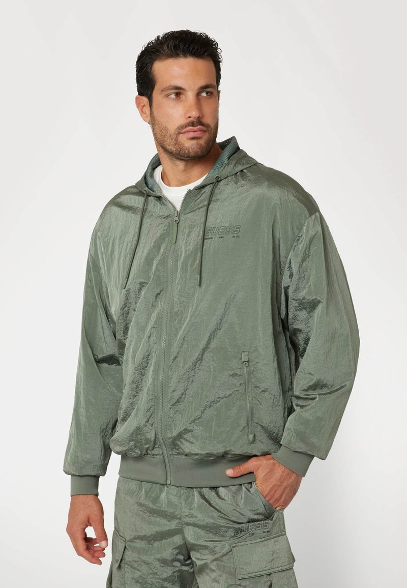 Vízálló zöld zipzáros kapucnis pulcsi texturált felülettel, húzózsinórral ellátott kapucnival és kenguru zsebekkel, összepárosítva megfelelő cargo sortokkal.